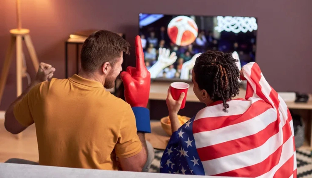 A couple watches a live sports game on TV, showing a man wearing oversized red foam gloves and a woman draped in an American flag, celebrating together with snacks and drinks.