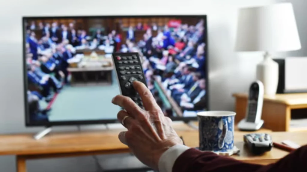 A smiling family with two young children sitting in a cozy living room, watching IPTV on a large smart TV. The screen displays a modern user interface with categories for live TV, movies, and shows. The atmosphere is warm, relaxed, and family-friendly.